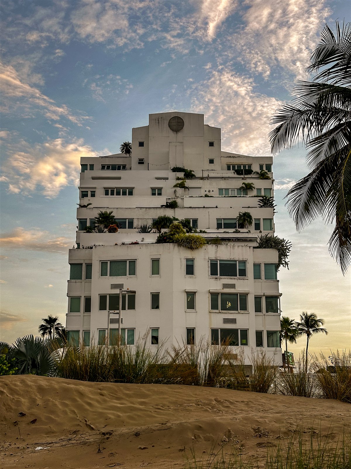 A white multi-story building above the sand at Isla Verde, San Juan, with palms and a cloudy evening sky.