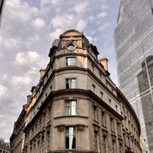 Looking up at an ornate old stone corner building with a domed turret beside a glass skyscraper in London.